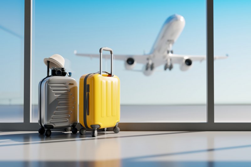 Silver suitcase with hat on atop and yellow suitcase side by side at airplane gate window with plane flying in background